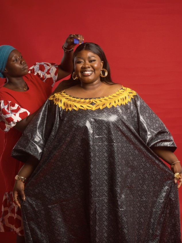 A smiling woman in a charcoal bazin grand boubou with gold embroidery while a tailor adjusts the fit