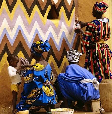 Women in wax print painting a zigzag mural on an earthen wall