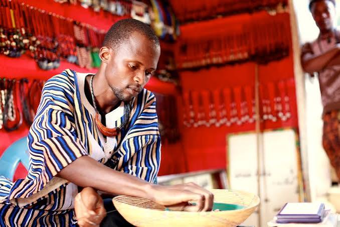 An artisan in a striped tunic working in a bead-filled market stall