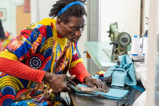 A tailor in colorful wax print cutting blue fabric with shears